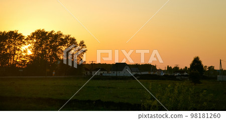 The setting sun behind the silhouettes of trees and the yellow evening sky over the houses in the village 98181260