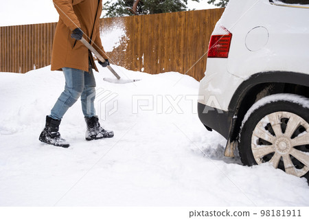 A man in winter cleans snow with a shovel in the yard of a house in the parking lot. Snowfall, difficult weather conditions, the car is stalling, digging up the passage 98181911