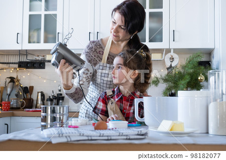 Mom and daughter in the white kitchen knead the dough in the bowl for gingerbread and cookies with a blender for Christmas and new year. 98182077