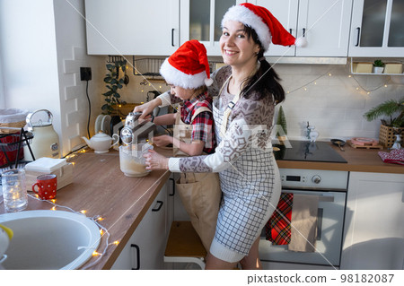Mom and daughter in the white kitchen knead the dough in the bowl for gingerbread and cookies with a blender for Christmas and new year. Mom and daughter in the white kitchen knead the dough in the bowl for gingerbread and cookies with a blender for Christmas and new year. 98182087