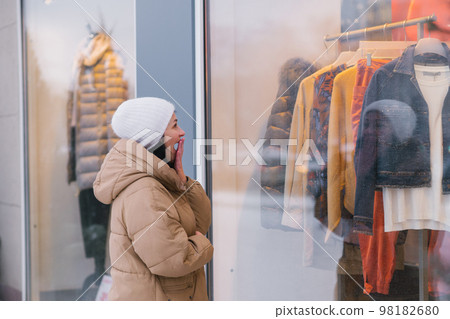 A woman in a white hat and coat covers her mouth with her hand and looks at the shop window A woman in a white hat and coat covers her mouth with her hand and looks at the shop window 98182680