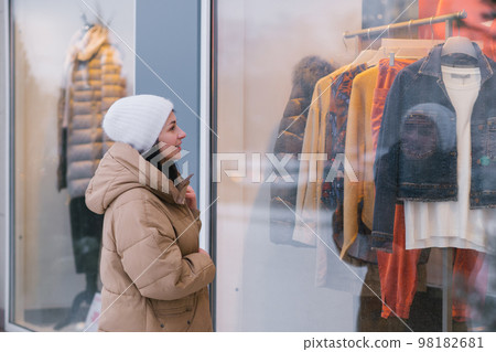 A stylish young woman in a hat and coat in the city looks into the windows of clothing stores A stylish young woman in a hat and coat in the city looks into the windows of clothing stores 98182681