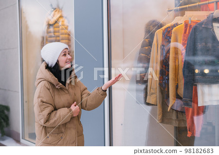 A stylish young woman in a hat and coat in the city looks into the windows of clothing stores A stylish young woman in a hat and coat in the city looks into the windows of clothing stores 98182683