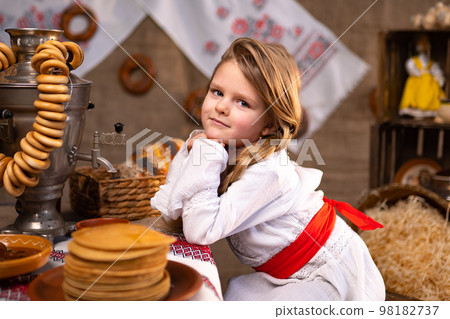 Cute little girl in folk costume seating at the table with samovar while celebrating Maslenitsa 98182737