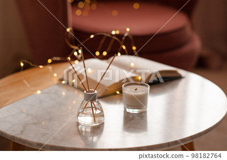 Cozy home atmosphere with liquid fragrance in glass bottle and sticks with open paper book over glow lights on marble table in living room close up. Cozy home atmosphere with liquid fragrance in glass bottle and sticks with open paper book over glow lights on marble table in living room close up. 98182764