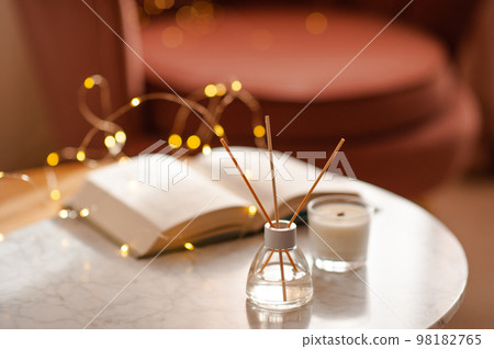 Cozy home atmosphere with liquid fragrance in glass bottle and sticks with open paper book over glow lights on marble table in living room close up. 98182765