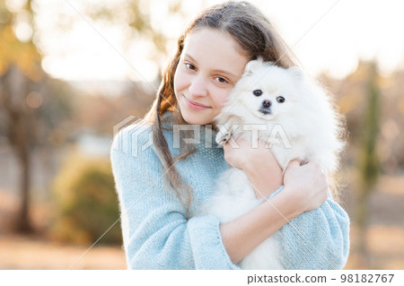 Cute smiling teenage girl 15-16 year old holding white fluffy spitz puppy dog together over autumn yellow nature background in park. Cute smiling teenage girl 15-16 year old holding white fluffy spitz puppy dog together over autumn yellow nature background in park. 98182767