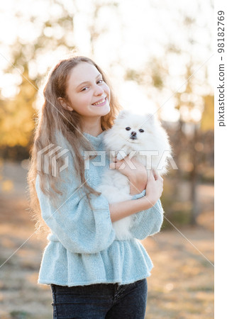 Cute smiling teenage girl 15-16 year old holding white fluffy spitz puppy dog together over autumn yellow nature background in park. 98182769