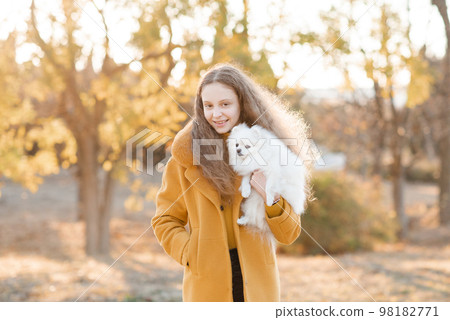 Cute smiling teenage girl 15-16 year old holding white fluffy spitz puppy dog together over autumn yellow nature background in park. Friendship. Cute smiling teenage girl 15-16 year old holding white fluffy spitz puppy dog together over autumn yellow nature background in park. Friendship. 98182771
