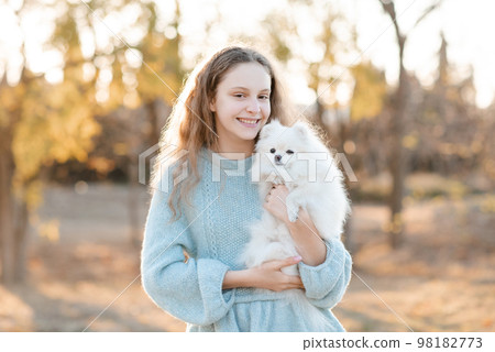 Cute smiling teenage girl 15-16 year old holding white fluffy spitz puppy dog together over autumn yellow nature background in park. Friendship. 98182773