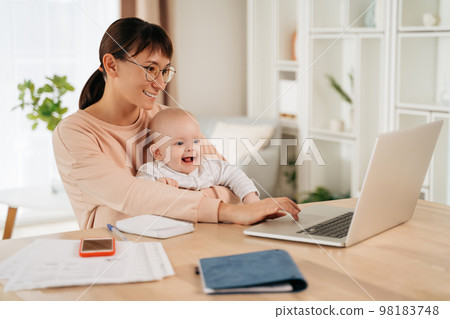 Young mother working online in home office with laptop and her son 98183748