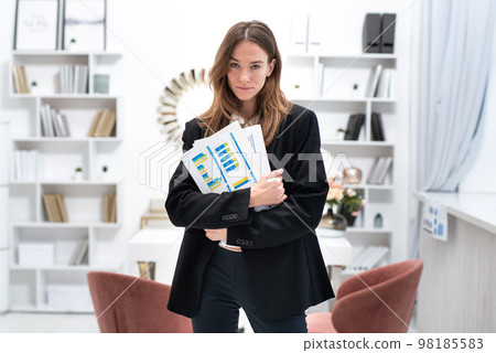 Portrait of a young woman standing in an office with documents Portrait of a young woman standing in an office with documents 98185583