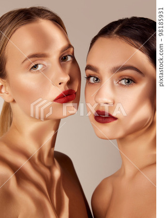 Natural look. Portrait of two young, beautiful girls with red lips makeup isolated over grey studio background. Shadows. Concept of skincare, cosmetology, natural beauty Natural look. Portrait of two young, beautiful girls with red lips makeup isolated over grey studio background. Shadows. Concept of skincare, cosmetology, natural beauty 98185931