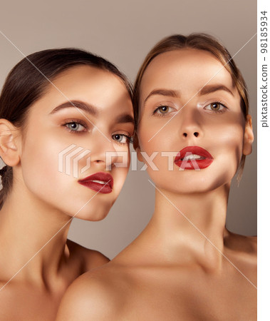 Perfect skin. Portrait of two young, beautiful girls with red lips makeup isolated over grey studio background. Shadows. Concept of skincare, cosmetology, natural beauty Perfect skin. Portrait of two young, beautiful girls with red lips makeup isolated over grey studio background. Shadows. Concept of skincare, cosmetology, natural beauty 98185934