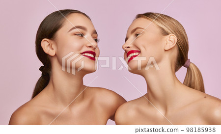 Red lipstick beauty. Portrait of two young, beautiful girls with well-kept skin isolated over pink studio background. Concept of skincare, cosmetology, natural beauty Red lipstick beauty. Portrait of two young, beautiful girls with well-kept skin isolated over pink studio background. Concept of skincare, cosmetology, natural beauty 98185938