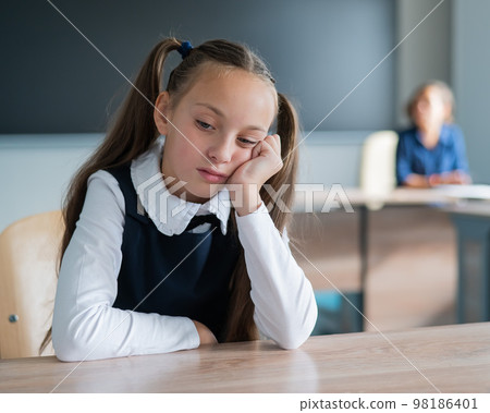 Little caucasian girl is bored at the lesson at school. The schoolgirl is sitting at her desk and the teacher is sitting in the background. 98186401