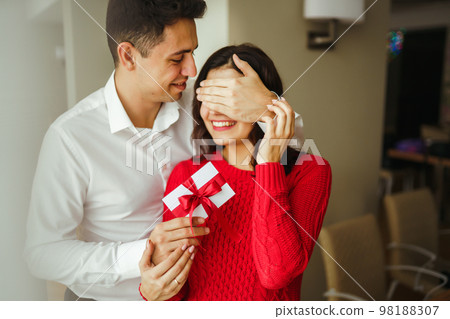A young man gives his beloved a present and closes her eyes with a hand. Valentine's Day. 98188307
