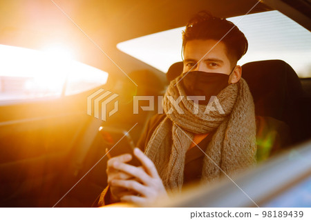 Young man in protective sterile mask in taxi car on a back seat checking her cell phone. Young man in protective sterile mask in taxi car on a back seat checking her cell phone. 98189439