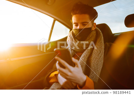 Young man in protective sterile mask in taxi car on a back seat checking her cell phone. Young man in protective sterile mask in taxi car on a back seat checking her cell phone. 98189441