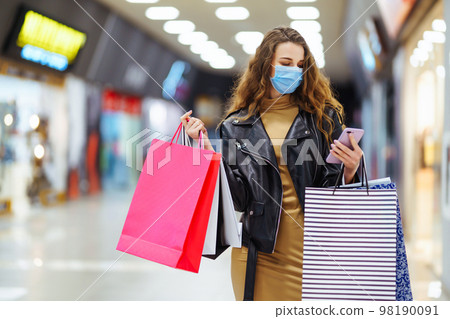 Young woman in protective mask using mobile phone while holding shopping bag in department store. 98190091