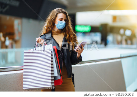 Fashionable woman in protective medical mask with multi-colored bags in modern shop center. Fashionable woman in protective medical mask with multi-colored bags in modern shop center. 98190108