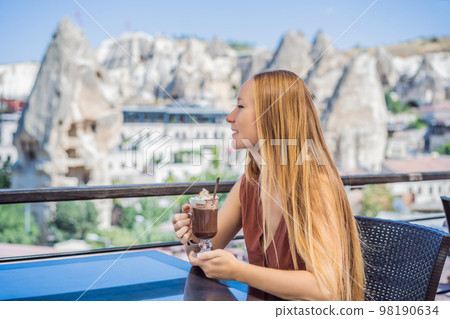 Young woman drinking coffee on the background valley with rock formations and fairy caves near Goreme in Cappadocia Turkey 98190634