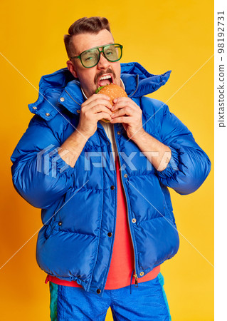 Delicious. Portrait of handsome man in blue winter jacket posing with burger over bright yellow background. Concept of leisure time, winter holiday, hobby 98192731