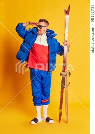 Portrait of handsome man in blue winter jacket and slippers posing with skis over bright yellow background. Concept of leisure time, winter sport, hobby 98192735
