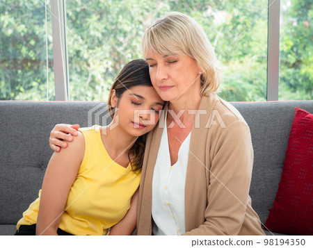 The adopted caucasian mother and Asian teen daughter happily hugged and smiled on the sofa. love and sharing time of stepmother, stepdaughter concept 98194350