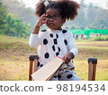 A cute little African-American girl in eyeglasses is sitting on a chair and holding a notepad and a pencil in a public Park on a beautiful morning.. 98194534
