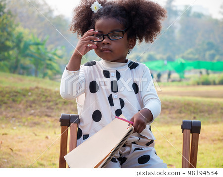 A cute little African-American girl in eyeglasses is sitting on a chair and holding a notepad and a pencil in a public Park on a beautiful morning.. A cute little African-American girl in eyeglasses is sitting on a chair and holding a notepad and a pencil in a public Park on a beautiful morning.. 98194534