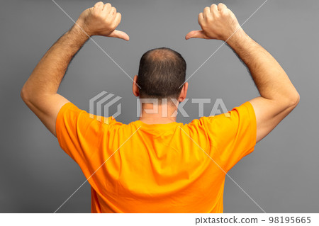 Proud man in orange t-shirt with his back turned pointing with his thumbs at his receding bald spot, isolated on gray background. 98195665