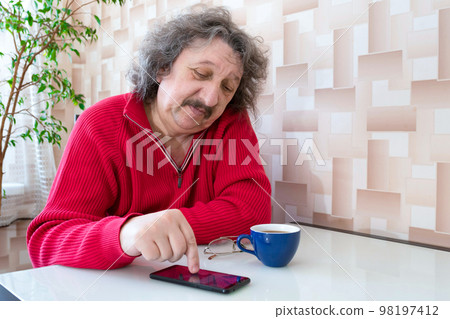 An elderly 60 or 70-year old man is sitting at the table with a cup of coffee and a phone in his hand An elderly 60 or 70-year old man is sitting at the table with a cup of coffee and a phone in his hand 98197412