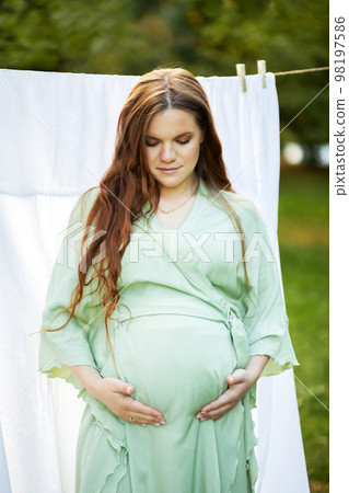 White pretty pregnant woman stands putting hands on big belly. Green trees, park,white sheet on background. Young healthy female wears dress. Baby shower,child delivery,preparation concept.Vertical  98197586
