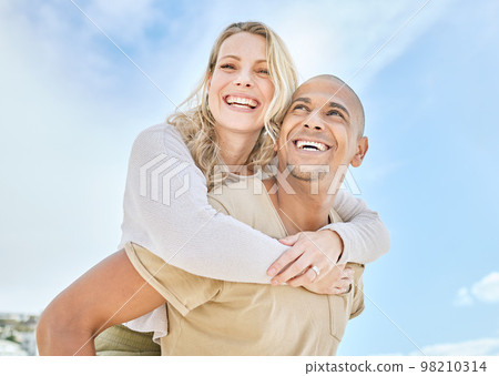 Woman, interracial couple and piggy back with love, happiness and fun in the sun outdoor. Happy people relationship with a smile in the sunshine together on travel and summer vacation with a blue sky 98210314