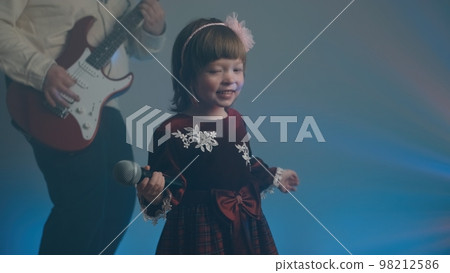 Little girl on stage in vintage dress, she sings into microphone and dances, her father plays an electric guitar. Color music is shining and smoke is billowing. Family time, creativity and hobbies Little girl on stage in vintage dress, she sings into microphone and dances, her father plays an electric guitar. Color music is shining and smoke is billowing. Family time, creativity and hobbies 98212586