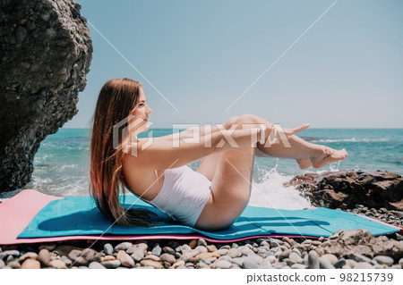 Woman yoga sea. Young woman with long hair in white swimsuit and boho style braclets practicing outdoors on yoga mat by the ocean on sunny day. Women's yoga fitness routine. Healthy lifestyle, harmony 98215739