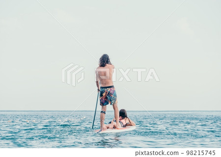 Sea woman and man on sup. Silhouette of happy young woman and man, surfing on SUP board, confident paddling through water surface. Idyllic sunset. Active lifestyle at sea or river. 98215745