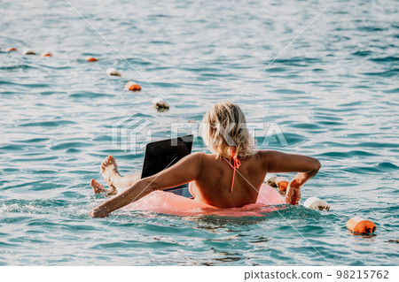 Woman works on laptop in sea. Freelancer, young blond woman in sunglases floating on an inflatable big pink donut with a laptop in the sea at sunset. Freelance, travel and holidays concept. 98215762