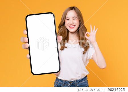 Cheerful beautiful Asian woman holding smartphone and shows ok sign on light yellow background. Cheerful beautiful Asian woman holding smartphone and shows ok sign on light yellow background. 98216025