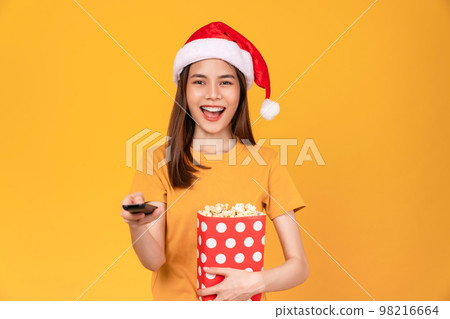 woman wearing Christmas hat with holding a red bucket of popcorn while the TV remote to watch movies 98216664