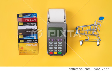 Stack of credit cards and card reader machine with shopping basket on yellow background.  98217336