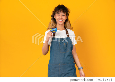 Cheerful beautiful Asian woman in white t-shirt and holding mockup credit card on yellow background. Cheerful beautiful Asian woman in white t-shirt and holding mockup credit card on yellow background. 98217341