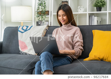 Cheerful young woman sitting on sofa and using laptop computer at home. 98217684