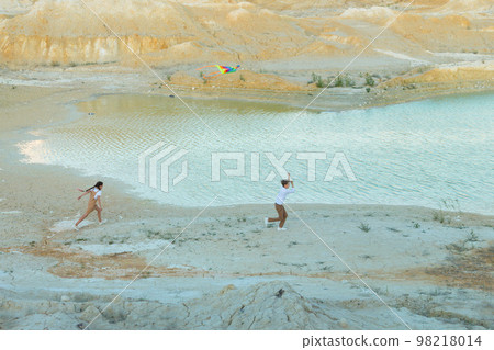 A boy and a girl with a kite are running through a mountainous area along a river 98218014