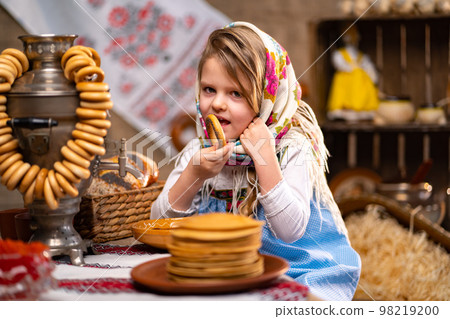 Little girl in folk Russian headscarf and dress eating bagels and celebrating Maslenitsa 98219200