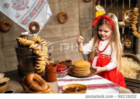 Little cute girl in folk costume sprinklers pancakes with honey while celebrating Shrovedite  98219202