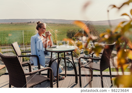 A middle-aged woman sits in a street cafe overlooking the mountains at sunset. She is dressed in a blue jacket and drinks coffee admiring the nature. Travel and vacation concept. 98223684