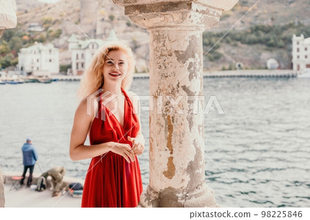 Redhead woman portrait. Curly redhead young caucasian woman with freckles looking at camera and smiling. Cute woman close up portrait in a red long dress posing on sea and travel city background Redhead woman portrait. Curly redhead young caucasian woman with freckles looking at camera and smiling. Cute woman close up portrait in a red long dress posing on sea and travel city background 98225846
