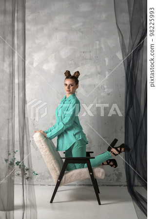 Girl in turquoise suit on grey wall background.Beautiful Young woman with bun hairstyle poses on chair in fashion studio 98225993